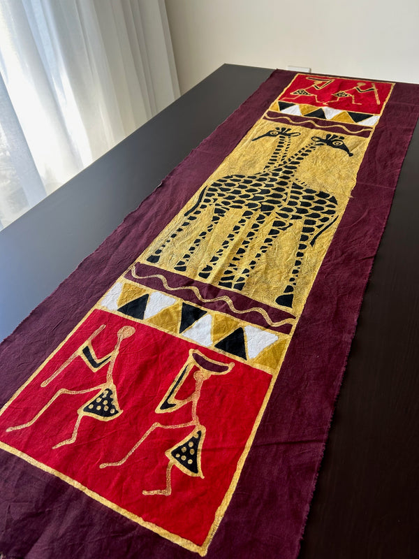 Batik table cloth featuring two giraffes crossing necks, hand-painted in vibrant red and complementary hues, displayed on a dining table.