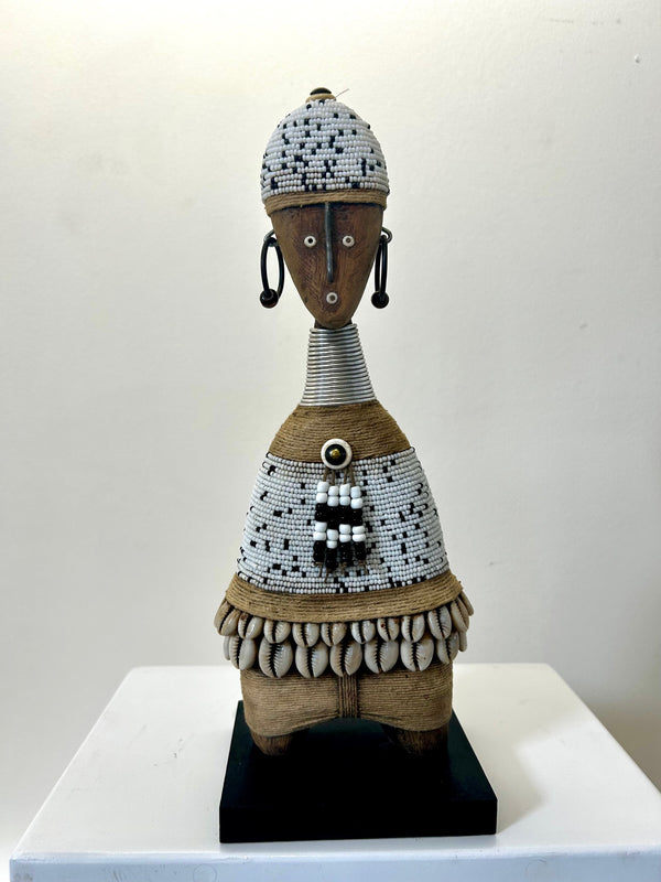 Beaded Namji doll from Cameroon, adorned with white and black beads and cowrie shells, representing fertility and motherhood, displayed on a pedestal.