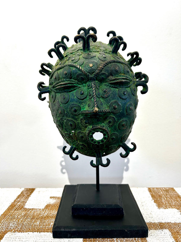 Bronze Ife tribal mask from Benin with intricate patterns and scarification marks, displayed on a stand at Rogl Souvenirs & Gallery, African art gallery.