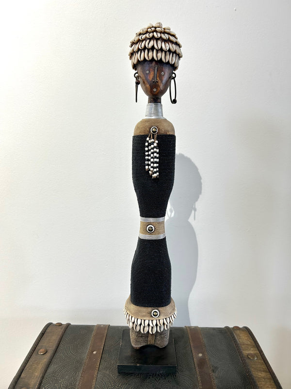 Beaded Namji doll from Cameroon, adorned with cowrie shells and intricate beads, representing fertility and motherhood, displayed on a wooden base.