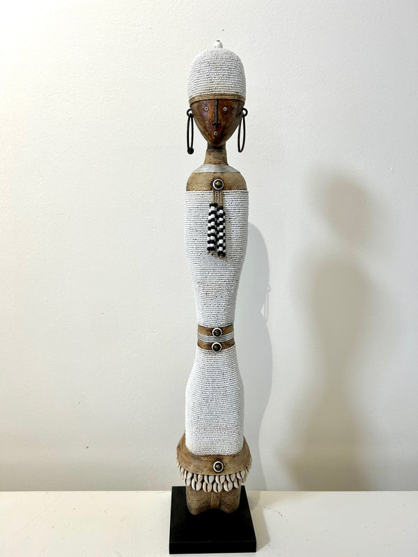 Beaded Namji doll from Cameroon, adorned with white beads and cowrie shells, representing fertility and motherhood, displayed at Rogl Souvenirs & Gallery.