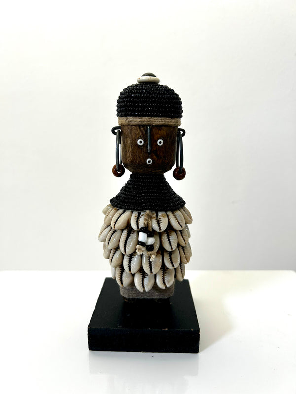 Beaded Namji doll from Cameroon, adorned with black beads and cowrie shells, representing fertility and motherhood, displayed on a black base.