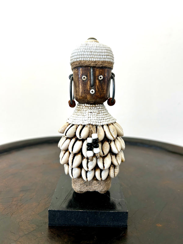 Namji doll sculpture from Cameroon, adorned with white beads and cowrie shells, representing fertility and motherhood, displayed on a dark base.