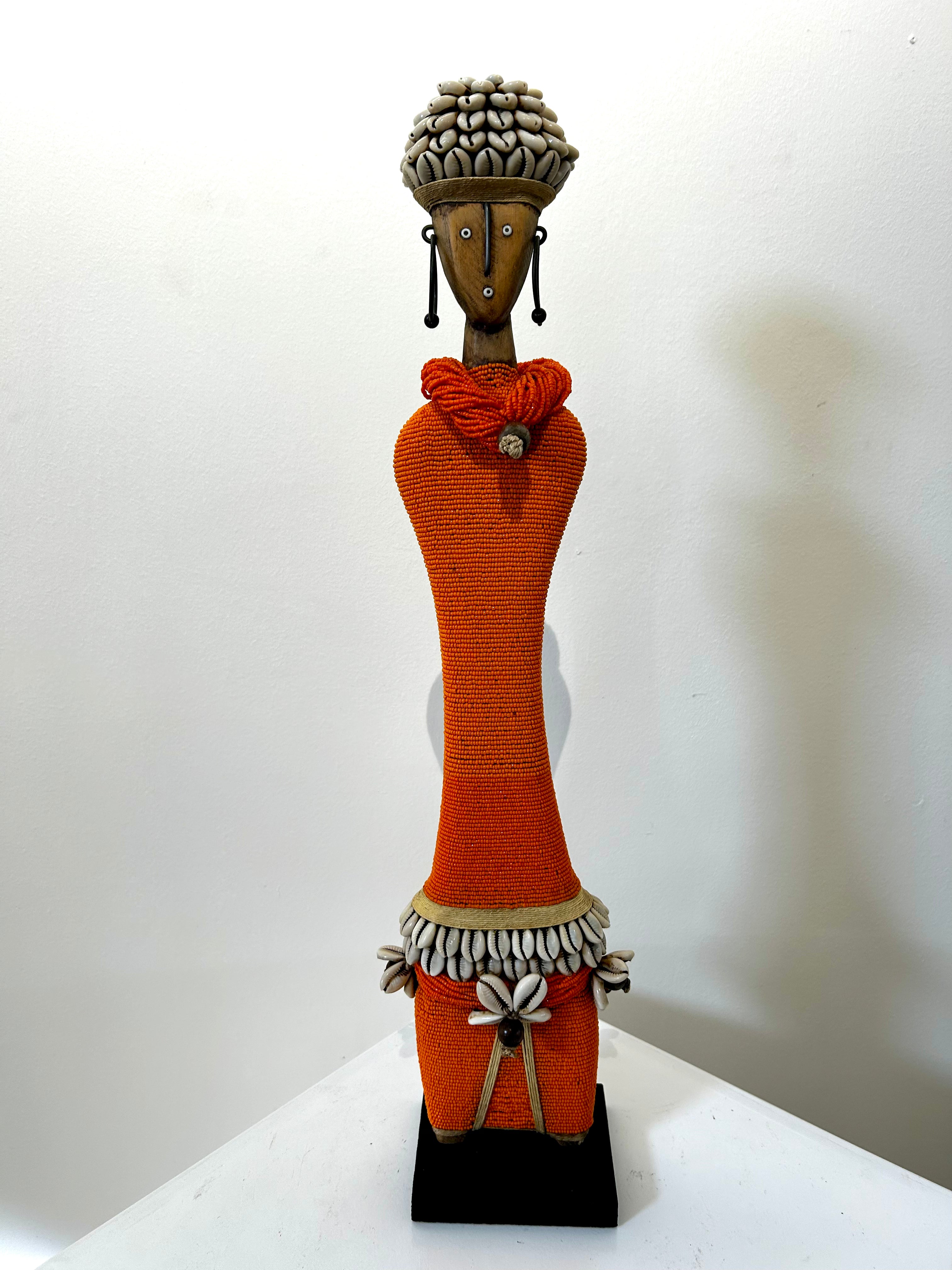 Orange Namji doll sculpture from Cameroon adorned with beads and cowrie shells, representing fertility and motherhood, displayed at Rogl Souvenirs & Gallery.