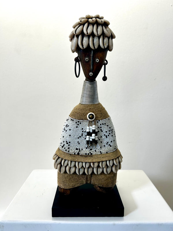 Namji doll sculpture from Cameroon adorned with beads and cowrie shells, representing fertility and motherhood, displayed at Rogl Souvenirs & Gallery.