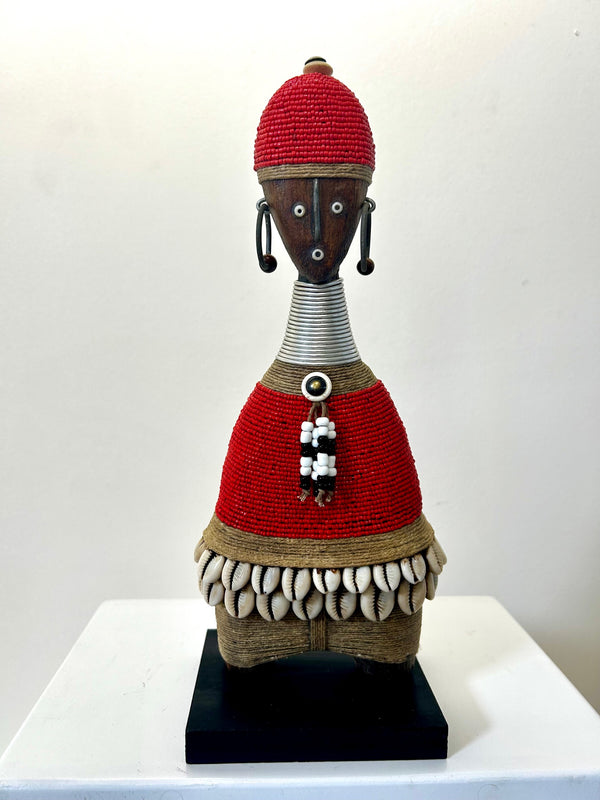 Namji doll sculpture from Cameroon, adorned with red beads and cowrie shells, representing fertility and motherhood, displayed at Rogl Souvenirs & Gallery.