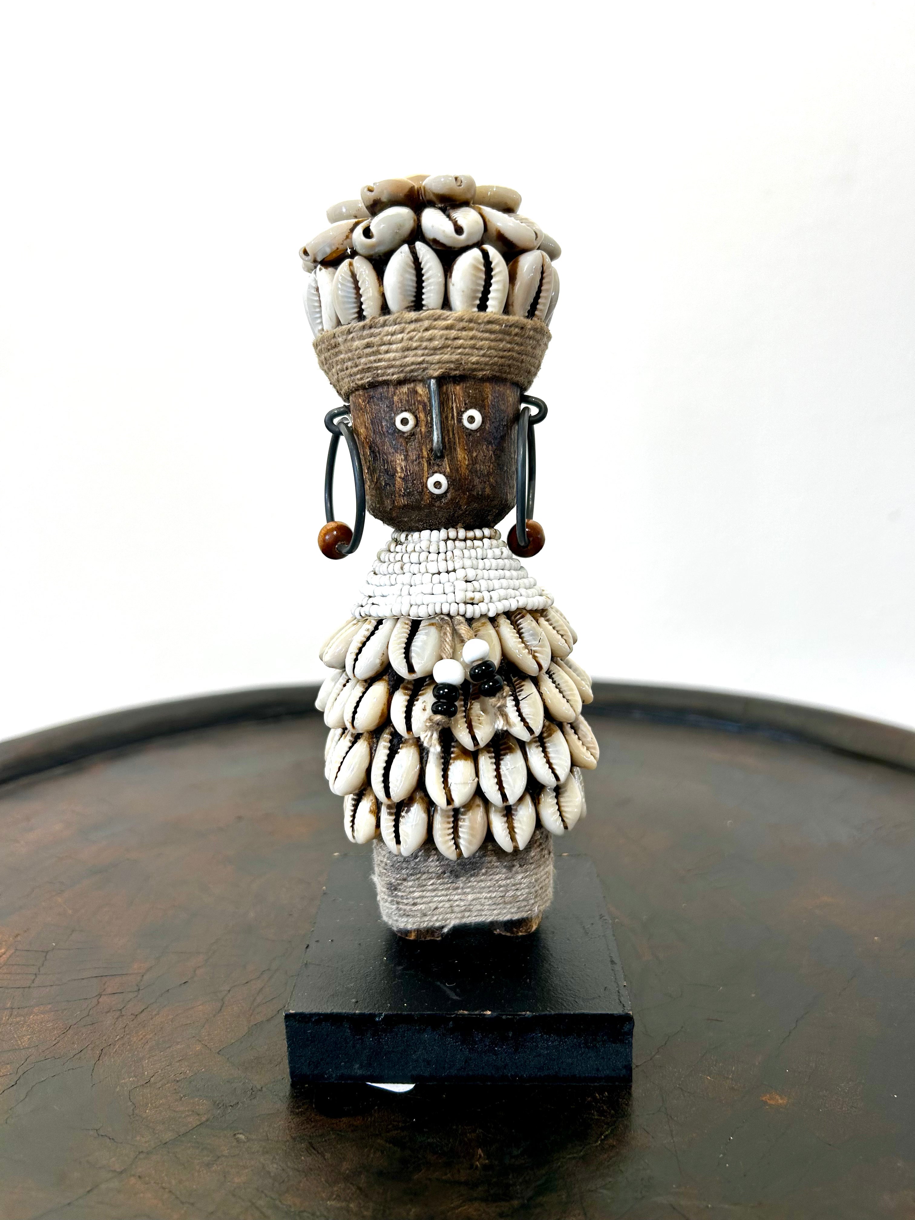 Namji doll sculpture from Cameroon adorned with white beads and cowrie shells, representing fertility and motherhood, displayed at Rogl Souvenirs & Gallery.