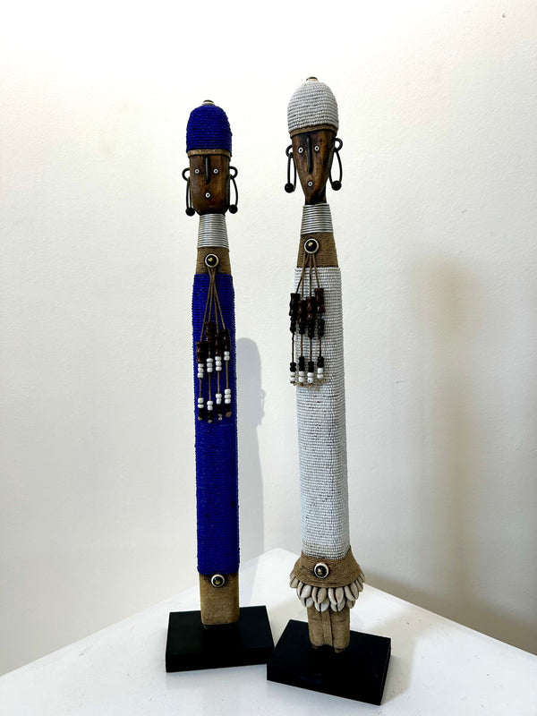 Indigo and white Namji dolls from Cameroon, adorned with beads and cowrie shells, representing fertility and motherhood, displayed at Rogl Souvenirs & Gallery.