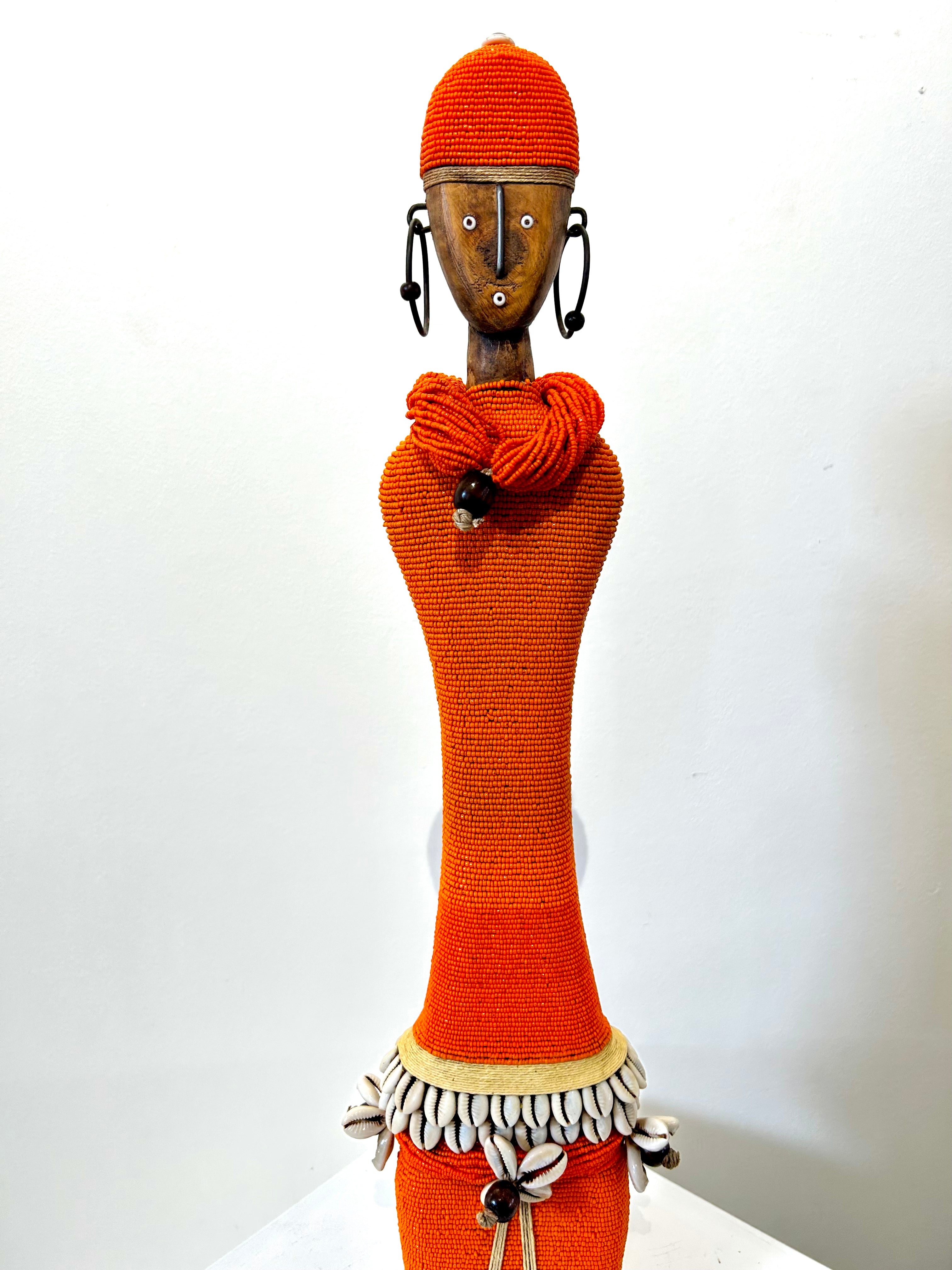 Orange beaded Namji doll from Cameroon adorned with cowrie shells, representing fertility and motherhood, displayed at Rogl Souvenirs & Gallery.