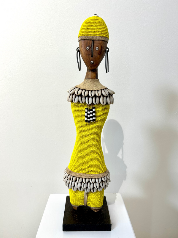Yellow beaded Namji doll from Cameroon adorned with cowrie shells, representing fertility and motherhood, displayed at Rogl Souvenirs & Gallery.