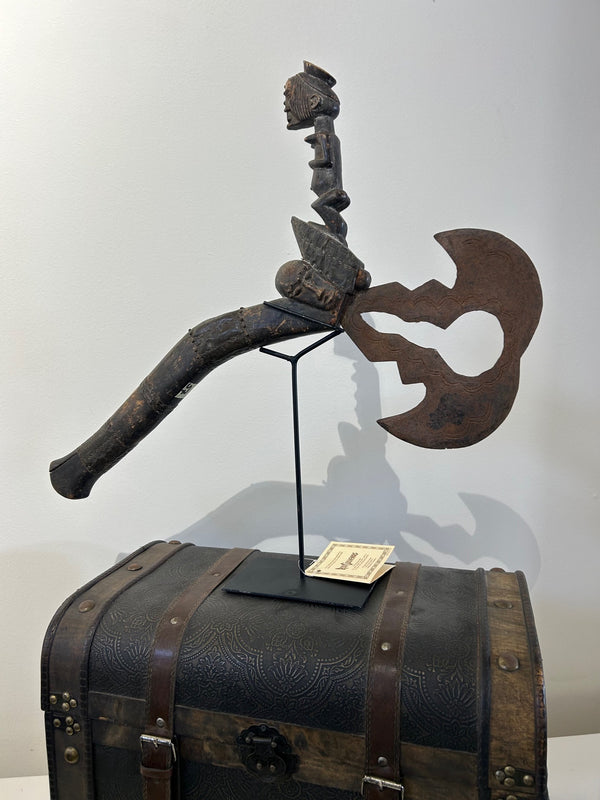 Antique Fang metal hatchet from Congo-Zaire with carved wooden handle and patina, displayed on a stand at Rogl Souvenirs & Gallery.