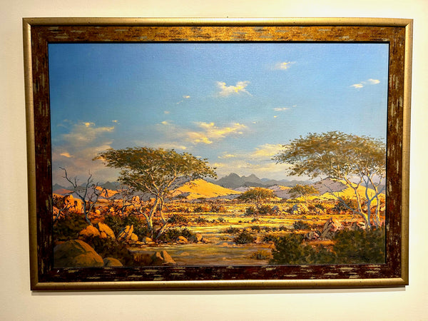 Breathtaking landscape painting of Damaraland by Namibian artist Archie van der Ploeg, featuring majestic mountains and rugged terrains under a blue sky, displayed in a gallery frame.