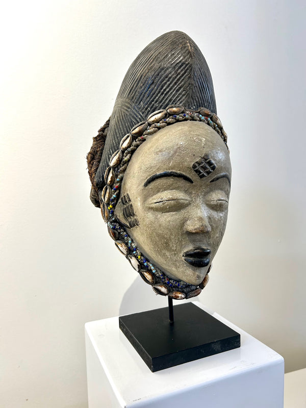 Ceremonial Punu mask from Gabon featuring intricate craftsmanship with cowrie shells, braided palm leaves, and colorful beads, displayed at Rogl Souvenirs & Gallery.