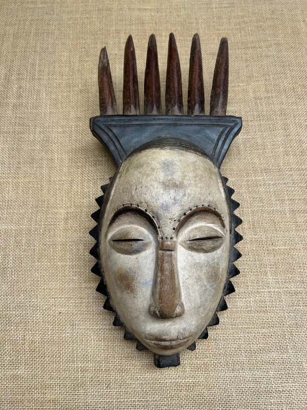 Traditional Igbo ceremonial mask from Nigeria with intricate carvings and regal headgear, featuring white and brown colors, displayed on a textured background.