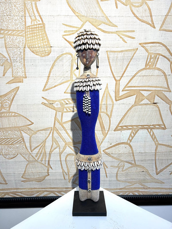 Beaded Namji doll in indigo with cowrie shells, handcrafted from pine wood, representing fertility and motherhood, displayed at Rogl Souvenirs & Gallery, African art gallery.