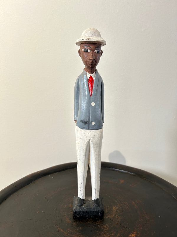 Wooden colonial figure sculpture from Ivory Coast depicting a civil servant, hand-carved with multi-color details, displayed on a dark wooden surface.