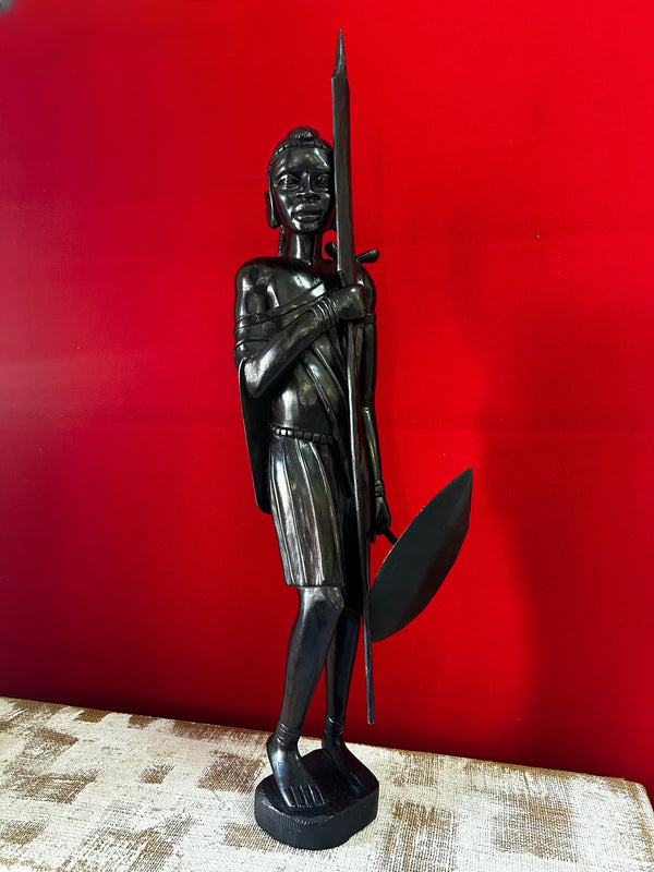 Ebony wood sculpture of a Maasai warrior from Tanzania, showcasing intricate craftsmanship and cultural essence, displayed against a red background at Rogl Souvenirs & Gallery.