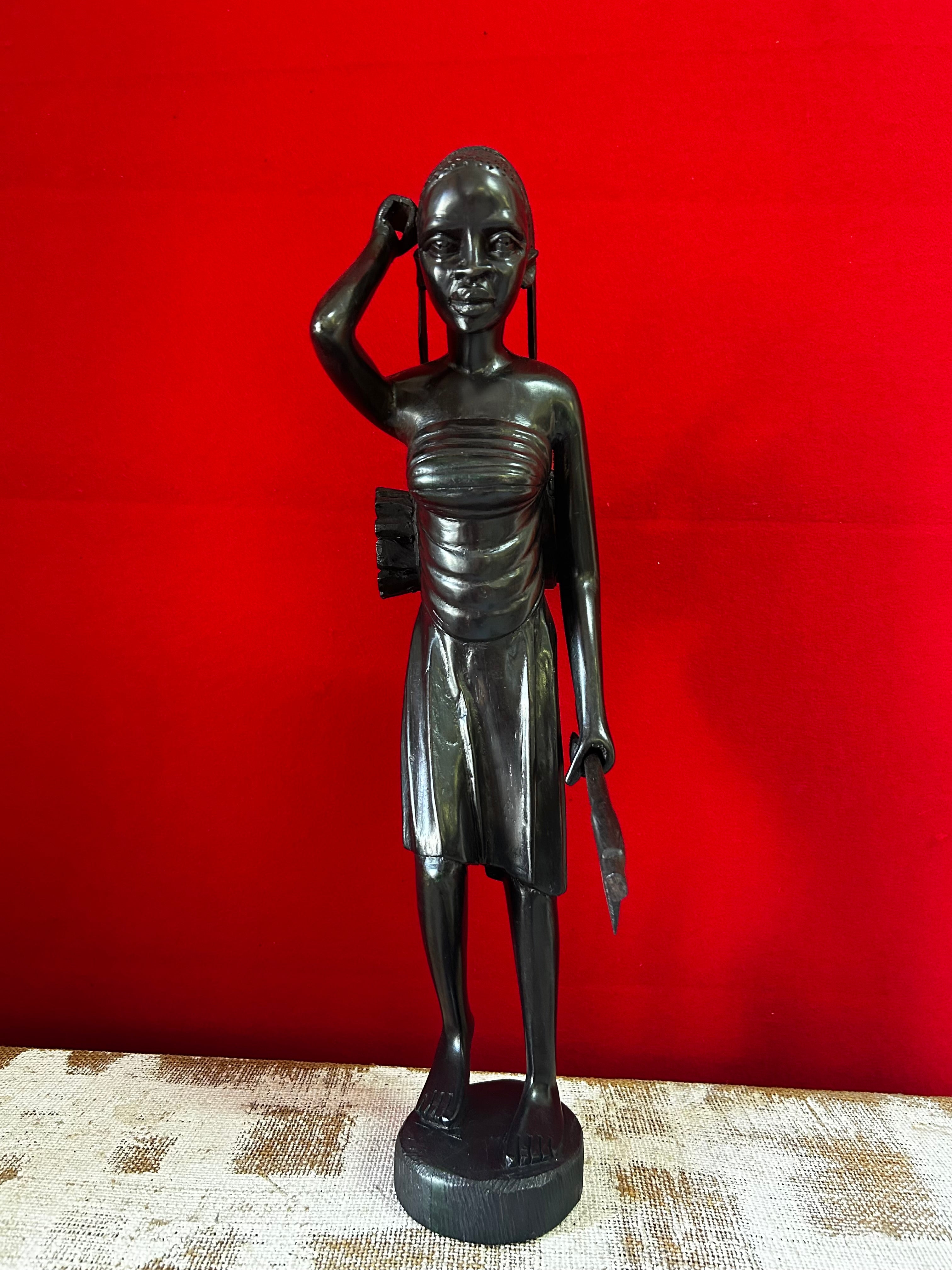 Sculpture of a Maasai woman gatherer in traditional attire, carved from ebony wood, displayed against a red background at Rogl Souvenirs & Gallery, African art gallery.