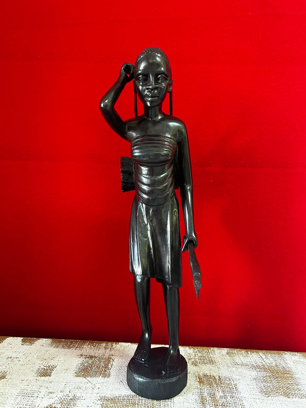 Sculpture of a Maasai woman gatherer in traditional attire, carved from ebony wood, displayed against a red background at Rogl Souvenirs & Gallery, African art gallery.
