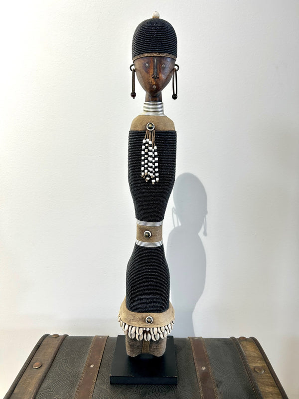 Beaded Namji doll in black with cowrie shells and intricate beadwork, representing fertility and motherhood from the Namji tribe, Cameroon, displayed on a wooden base.