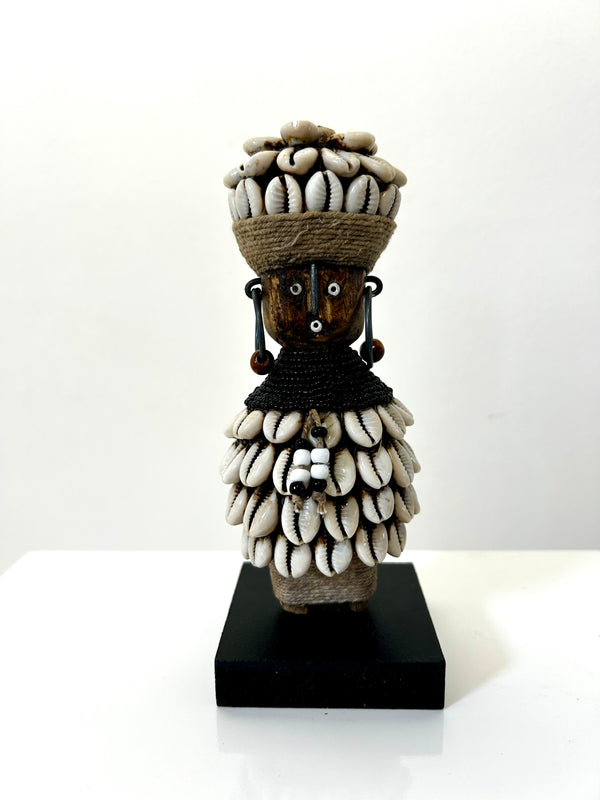 Beaded Namji doll from Cameroon, adorned with cowrie shells and beads, representing fertility and motherhood, displayed on a black base.