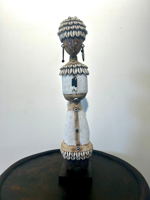 Beaded Namji doll from Cameroon adorned with cowrie shells and white beads, representing fertility and motherhood, displayed on a wooden base.