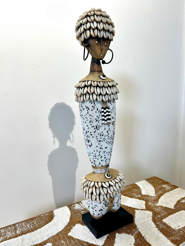 Beaded Namji doll in black and white, adorned with cowrie shells and intricate beads, representing fertility and motherhood from the Namji tribe of North Cameroon, displayed on a patterned surface.
