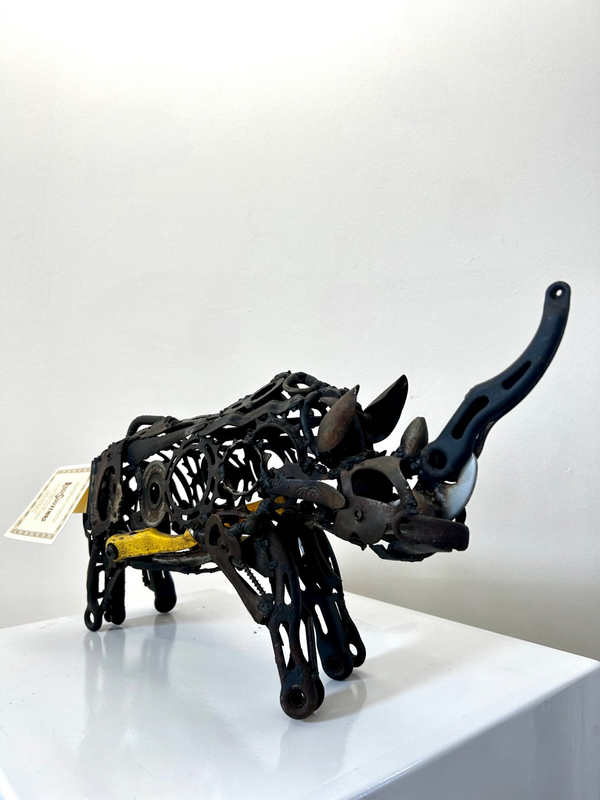 Sculpture of a rhino made from scrap metals, showcasing detailed craftsmanship and artistry, with a metallic grey finish, created by artist Ras from Namibia, displayed on a white surface.