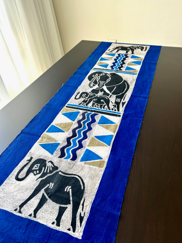 Batik style table cloth featuring a mother elephant and calf with intricate patterns, displayed on a dark wooden table, from Rogl Souvenirs & Gallery, African art gallery.