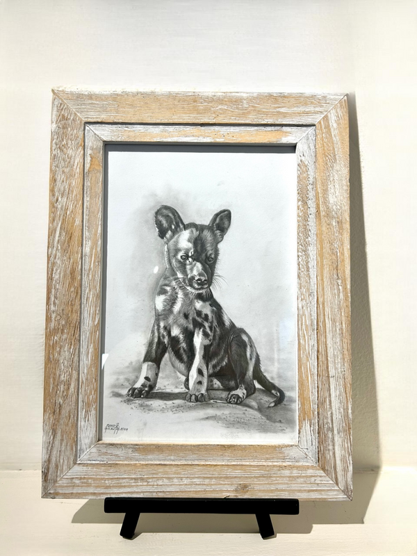 Pencil drawing of a sitting spotted wild dog puppy titled "Playful Innocence" by artist Moses, displayed in a rustic tabletop frame.